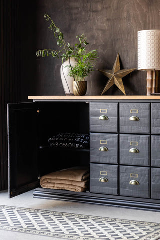 The Apothecary Sideboard with one door open with throws inside, styled in front of a dark wooden wall with various home accessories on the top.