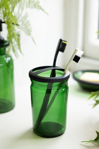 The Apothecary-Style Green Glass Toothbrush Holder styled with toothbrushes in, with matching glassware and plants in the background.