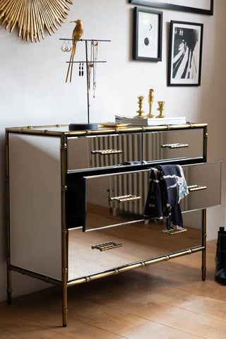 The Mirrored Bamboo Effect Chest Of Drawers styled in front of a wall with one drawer open, with various home accessories.