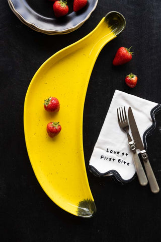 The Banana Serving Plate displayed on a black table with strawberries, some plates, a napkin and a knife and fork.
