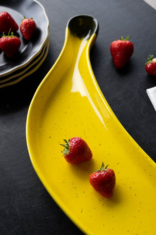 The Banana Serving Plate displayed on a black table with a stack of plates and some strawberries.