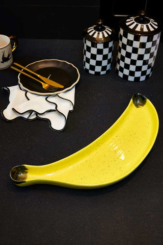 The Banana Serving Plate displayed on a black worktop with a mug, plate, cutlery, napkin and storage cannisters.