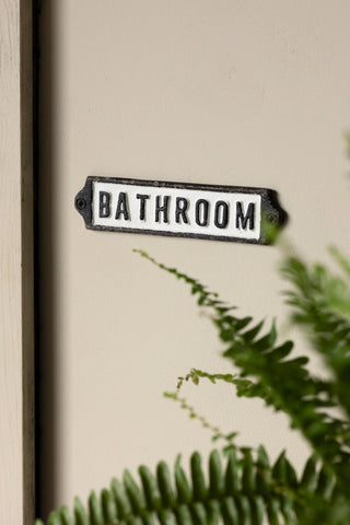 The Bathroom Door Hanging Sign on a neutral wall with a plant in the corner of the shot.