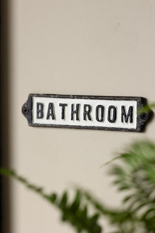 The Bathroom Door Hanging Sign on a neutral wall styled next to a plant.