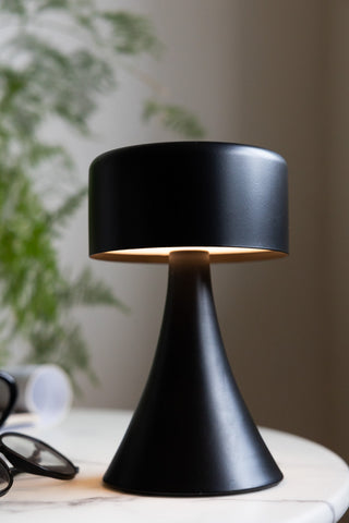 The Battery-Powered Cordless Table Lamp In Black switched on and displayed on a table with some accessories and a plant.