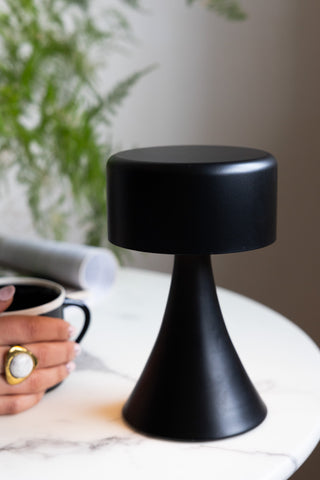 The Battery-Powered Cordless Table Lamp In Black on a round table, with someone's hand picking up a mug in the background.