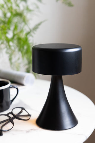 The Battery-Powered Cordless Table Lamp In Black styled on a table with some glasses, a mug, magazine and a plant.