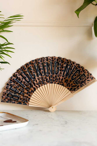 The Be Fucking Nice Leopard Print Fan on a white marble table displayed leaning against a wall, styled with a trinket tray and plants.