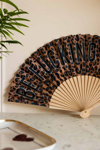 Close-up shot of the Be Fucking Nice Leopard Print Fan displayed on a white marble table leaning against the wall, next to a trinket tray and plant.