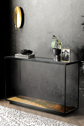 The Beautiful Burnished Metal & Glass Console Table with various accessories on, styled in front of a dark wall with a candle sconce on, with a plant and rug.