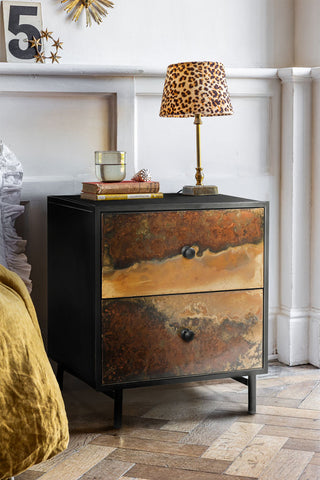 The Beautiful Burnished Metal Bedside Table styled with a table lamp, books, glass and shell on, next to a bed.