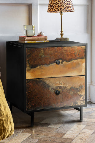 Close-up of the Beautiful Burnished Metal Bedside Table, styled with a lamp and various accessories on, next to a bed.