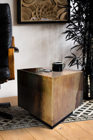 The Beautiful Burnished Metal Side Table styled with a mug and some glasses on, next to a chair with a plant and a framed artwork in the background.