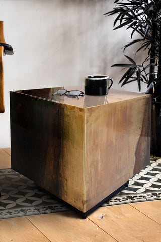 The Beautiful Burnished Metal Side Table with a mug and a pair of glasses on top,with a chair and a plant in the background.