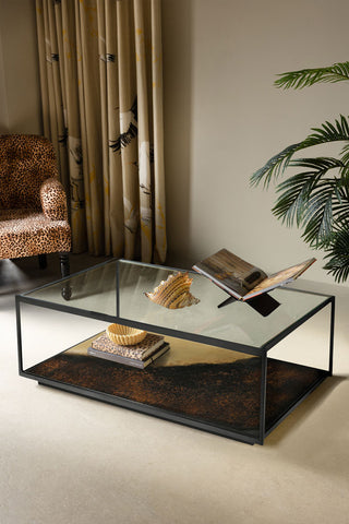 The Beautiful Burnished Metal & Glass Coffee Table styled with various accessories on, with an armchair, curtain and plant in the background.