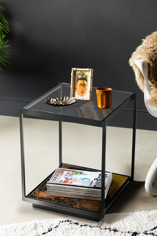 The Beautiful Burnished Metal & Glass Side Table styled with various accessories on, next to a rug, chair and plant.