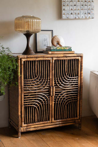 A statement dark brown rattan cabinet with an intricate symmetrical pattern on its doors, standing on four small legs, exuding natural elegance in a cosy interior.