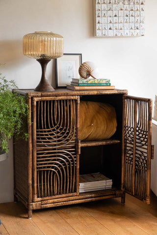 A beautifully woven rattan cabinet with two doors, finished in a deep brown tone, with organic curved detailing, styled with books, a glass lamp, and artwork.