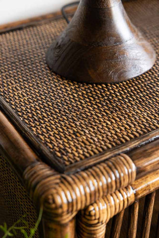 A vintage-style two-door rattan cabinet in a deep brown hue, showcasing a symmetrical curved design, placed on a wooden floor with a warm-toned lamp and books on top.