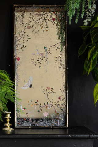 The Beautiful Pink Chinoiserie Decorative Mirror styled leaning against the wall on a black sideboard, with a candlestick holder and some greenery.