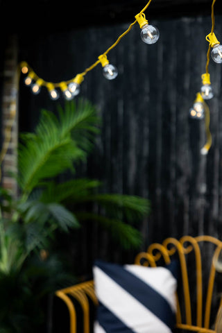 The Beautiful Yellow Festoon Lights draped across a garden with a garden bench, cushion, plant and fence in the background.
