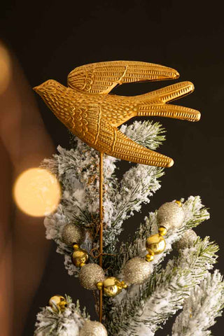 The Bird Christmas Tree Topper styled on the top of a Christmas tree, in front of a dark wall and glowing lights.