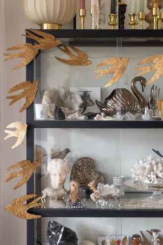 The Pretty Gold Birds Garland displayed draped around a black and glass cabinet full of decorative accessories.