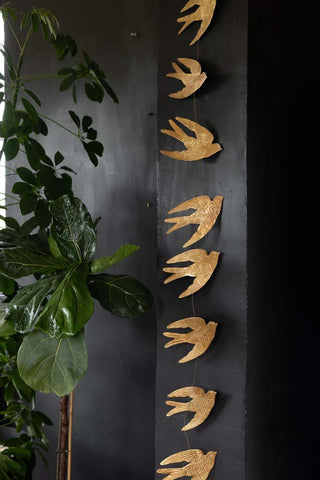 The Pretty Gold Birds Garland hanging in front of a black wall, styled next to a large plant.