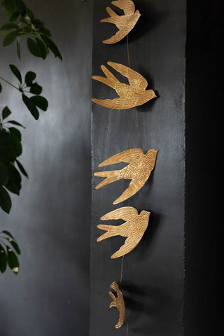 The Pretty Gold Birds Garland hanging vertically down a dark wall, next to some greenery.