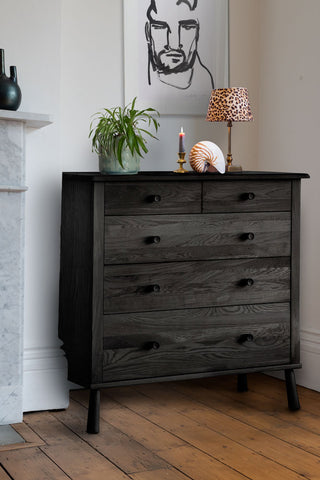 A lifestyle image of the Black 5-Drawer Chest styled with a plant, lamp, shell and candle, below an art print on the wall, next to a fireplace.