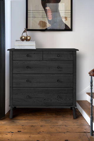 The Black 5-Drawer Chest styled with some magazines and a cherry ornament on, below an art print on the wall, next to a staircase.