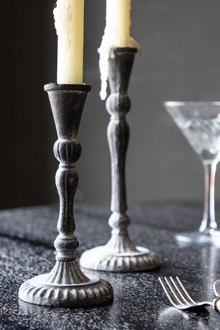 The Short Black Antique Zinc Style Candlestick Holder styled on a dining table with a candle inside, with the matching tall version in the background.