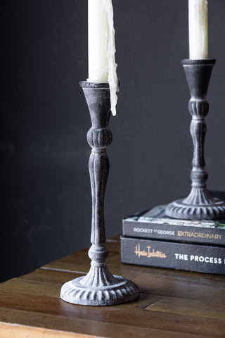 The Tall Black Antique Zinc Style Candlestick Holder styled on a wooden unit, with the short version on a stack of books in the background.