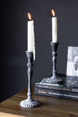 The Black Antique Zinc Style Candlestick Holders in Tall and Short, styled with lit candles on a wooden unit, with some books and a photograph.