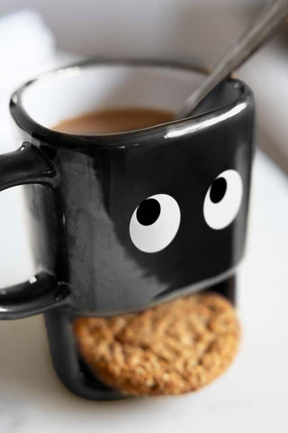 A black novelty mug with a face. The mug is filled with tea and there is a slot for biscuits