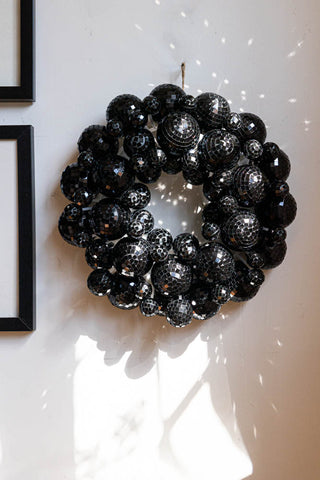 The Black Disco Ball Wreath displayed on a white wall, with the sun casting reflections around it, styled next to two framed art prints.