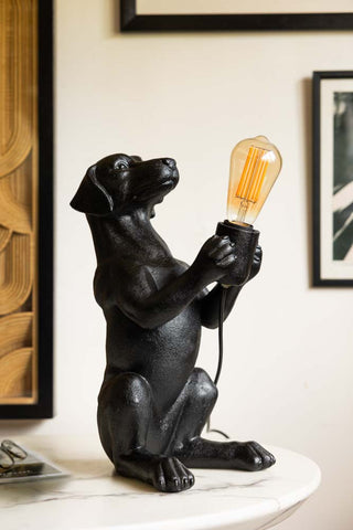 The Quirky Black Dog Lamp switched off, displayed on a table with a magazine and some glasses, with framed artwork on the wall in the background.