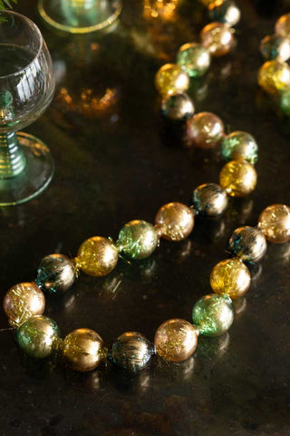 The Black, Green & Gold Bauble Garland styled on a dark surface with some glassware.