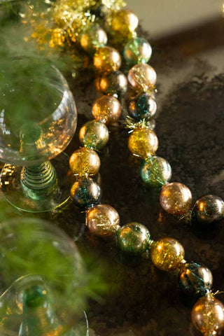 The Black, Green & Gold Bauble Garland styled on a dark surface with some greenery and a wine glass.