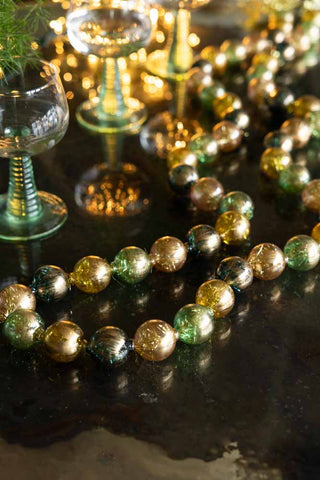 The Black, Green & Gold Bauble Garland styled on a dark surface amongst glassware and greenery.