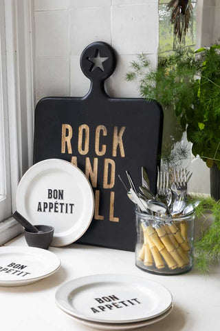 Lifestyle image showing the Black Rock And Roll Serving Board, Set Of 4 Bon Appetit White Bistro Side Plates and Beautiful 16-Piece Bamboo Design Cutlery Set, styled with a pestle and mortar and plants.