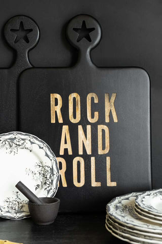 The Black Rock And Roll Serving Board leaning against a black wall, styled with the 12-Piece Vintage-Style Floral & Star Dinner Set and a black pestle and mortar.