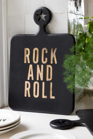 The Black Rock And Roll Serving Board displayed leaning against a tiled wall, styled with some plates and a plant.