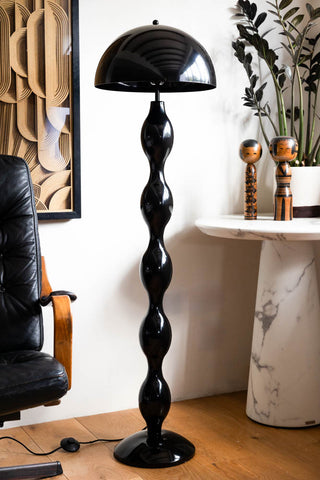 The Black Wavy Retro Floor Light displayed in a living room next to a swivel chair and a side table, styled with a potted plant, some ornaments and a framed artwork on the wall.