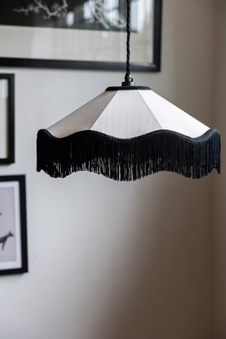 The Black & Cream Tassel Ceiling Light Shade hanging in front of a wall with black framed art prints on.