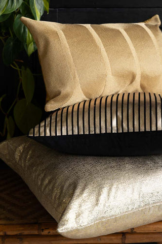 The Black & Gold Striped Cushion styled in a stack of cushions on a wicker bench, with some greenery in the background.