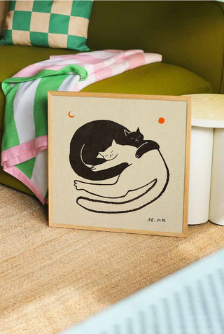 Two cuddle cats under an orange moon. The art print is framed and leaning against a green sofa.