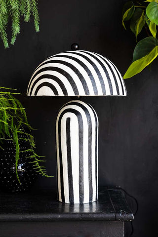 The Black & White Stripe Paper Mache Table Lamp displayed on a black sideboard with greenery.