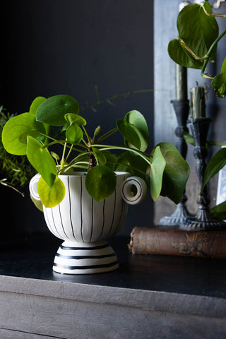 The Black & White Stripe Vase with Handles styled on a black surface with a plant inside, with other home accessories in the background.