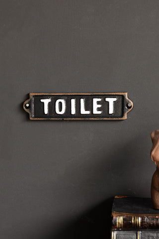 The Black & White Toilet Door Hanging Sign displayed on a black wall above a stack of books and a candle.